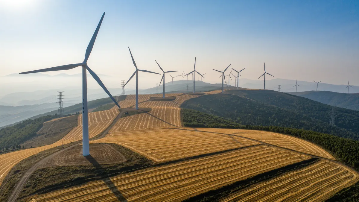 Windräder auf einem Höhenrücken über abgeernteten Feldern, dahinter Strommasten